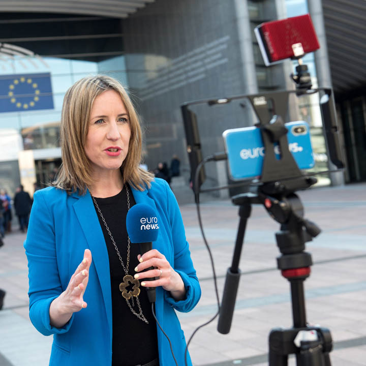 Kobieta reporterka kanału Euronews stoi przed smartfonem zamontowanym na statywie i nagrywa relację